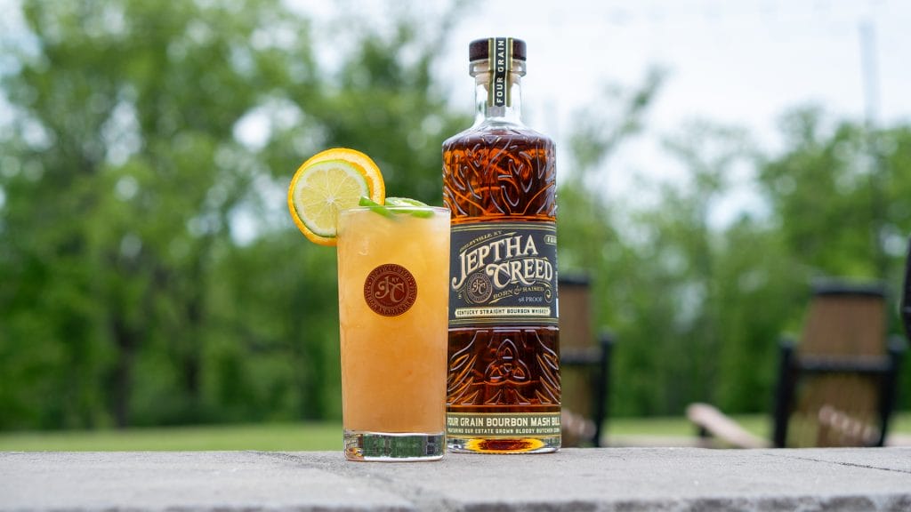 Cocktail and bottle of Jeptha Creed Four Grain on cement wall in front of spring landscape.
