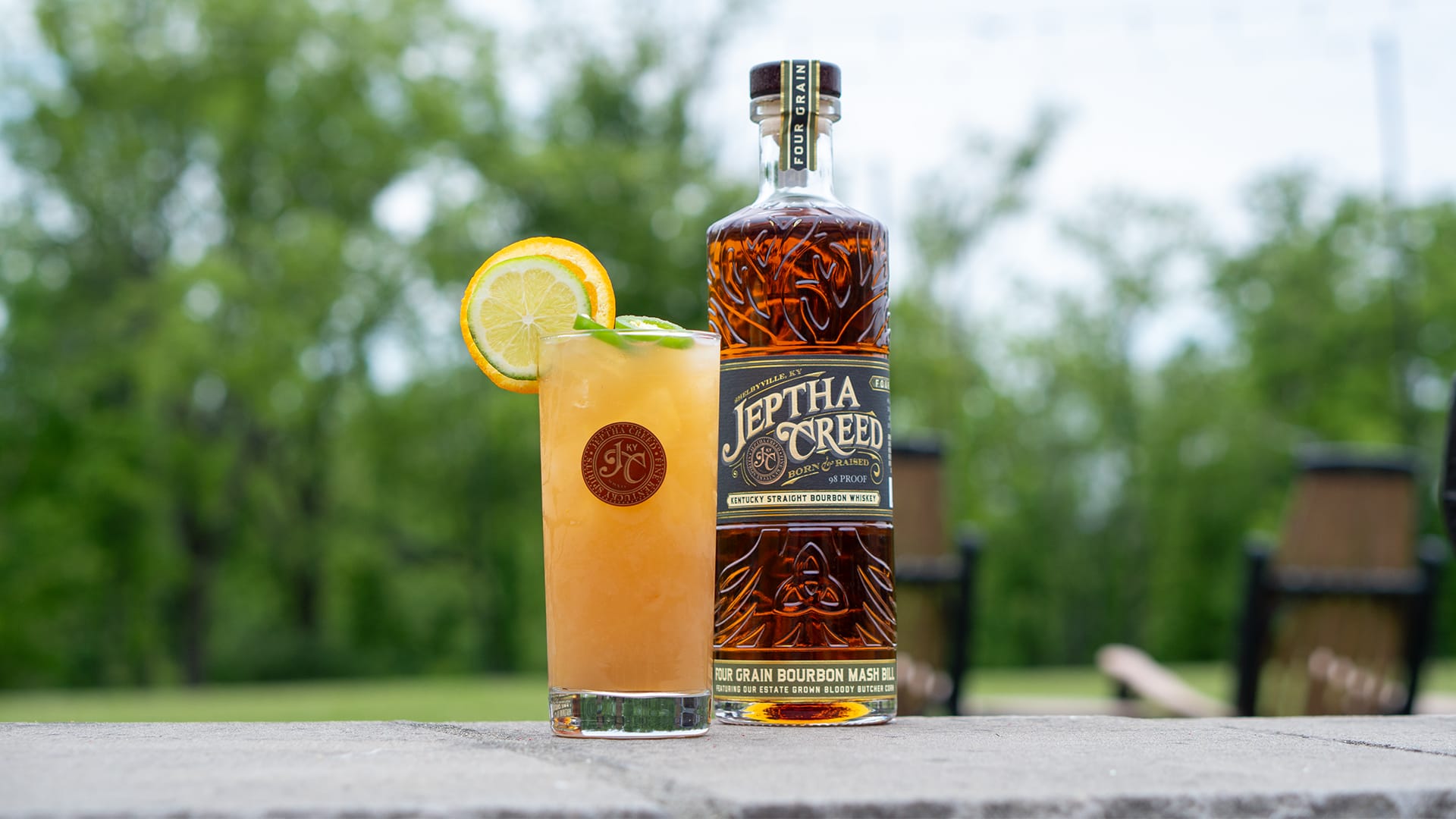 Cocktail and bottle of Jeptha Creed Four Grain on cement wall in front of spring landscape.
