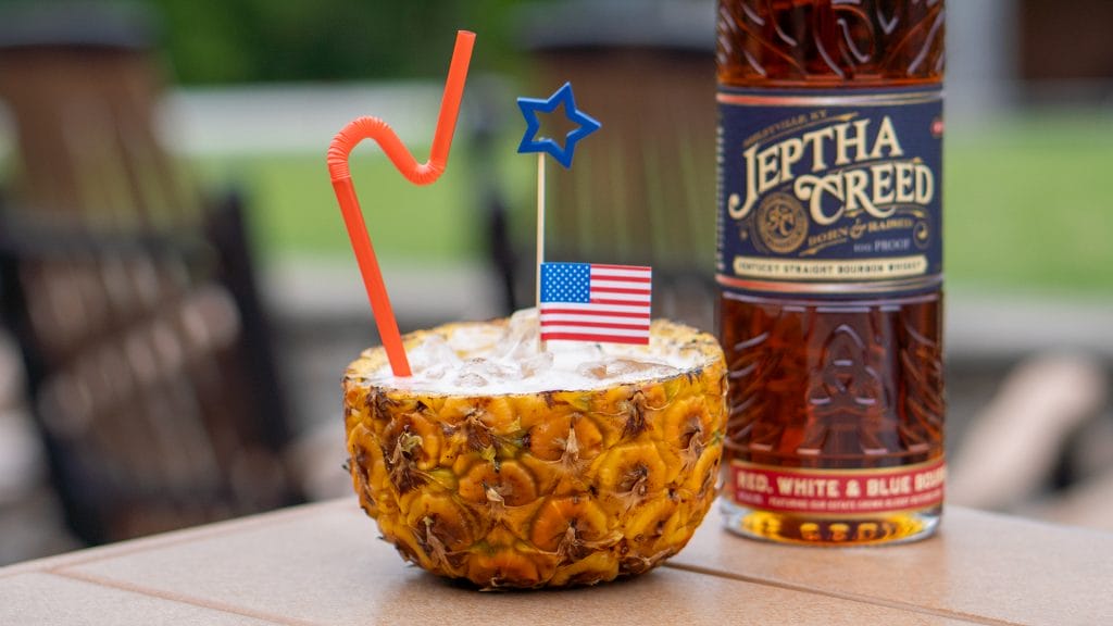Photo of cocktail inside of pineapple with Jeptha Creed Red, White & Blue bourbon sitting next to it on a table.
